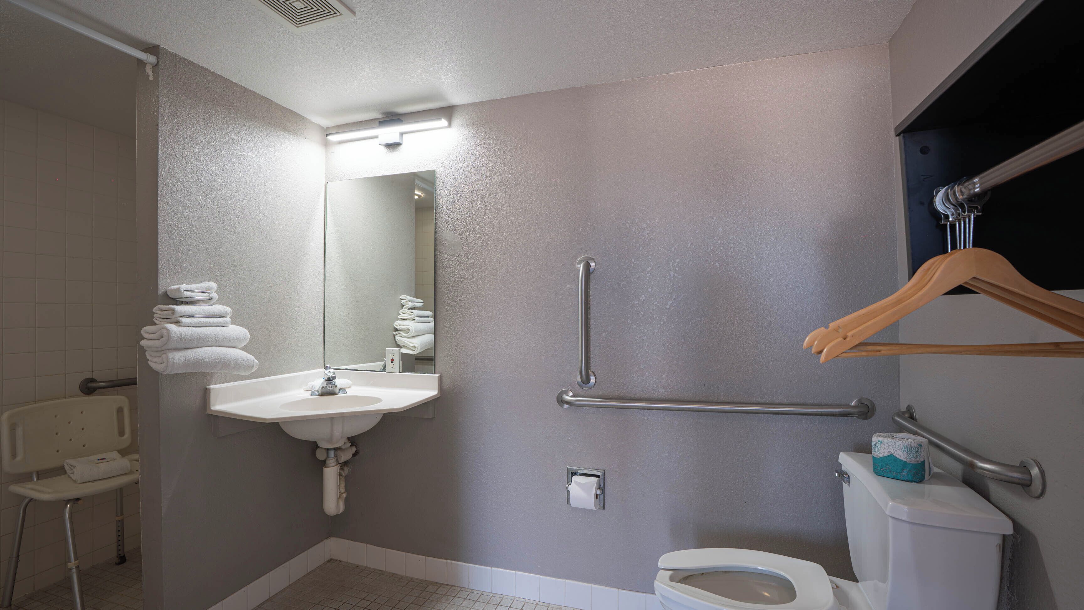A bathroom with a sink, mirror, toilet, grab bars, and a towel rack.