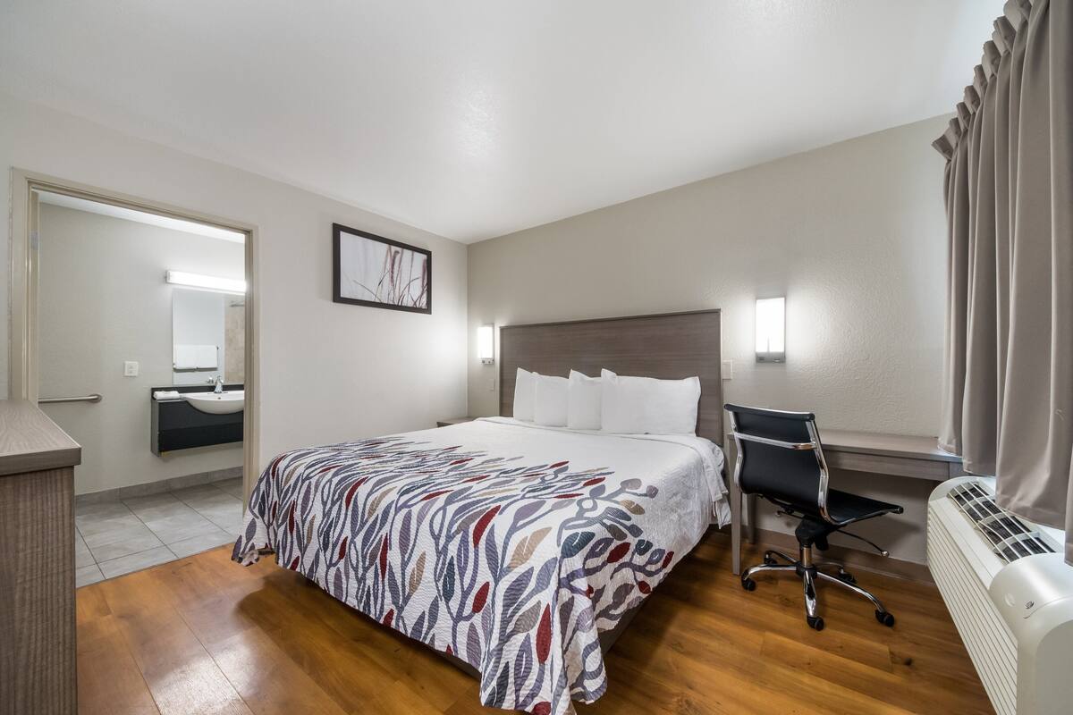 A hotel room with a bed, a desk, a chair, a bedside table, a mirror, and a window with curtains.