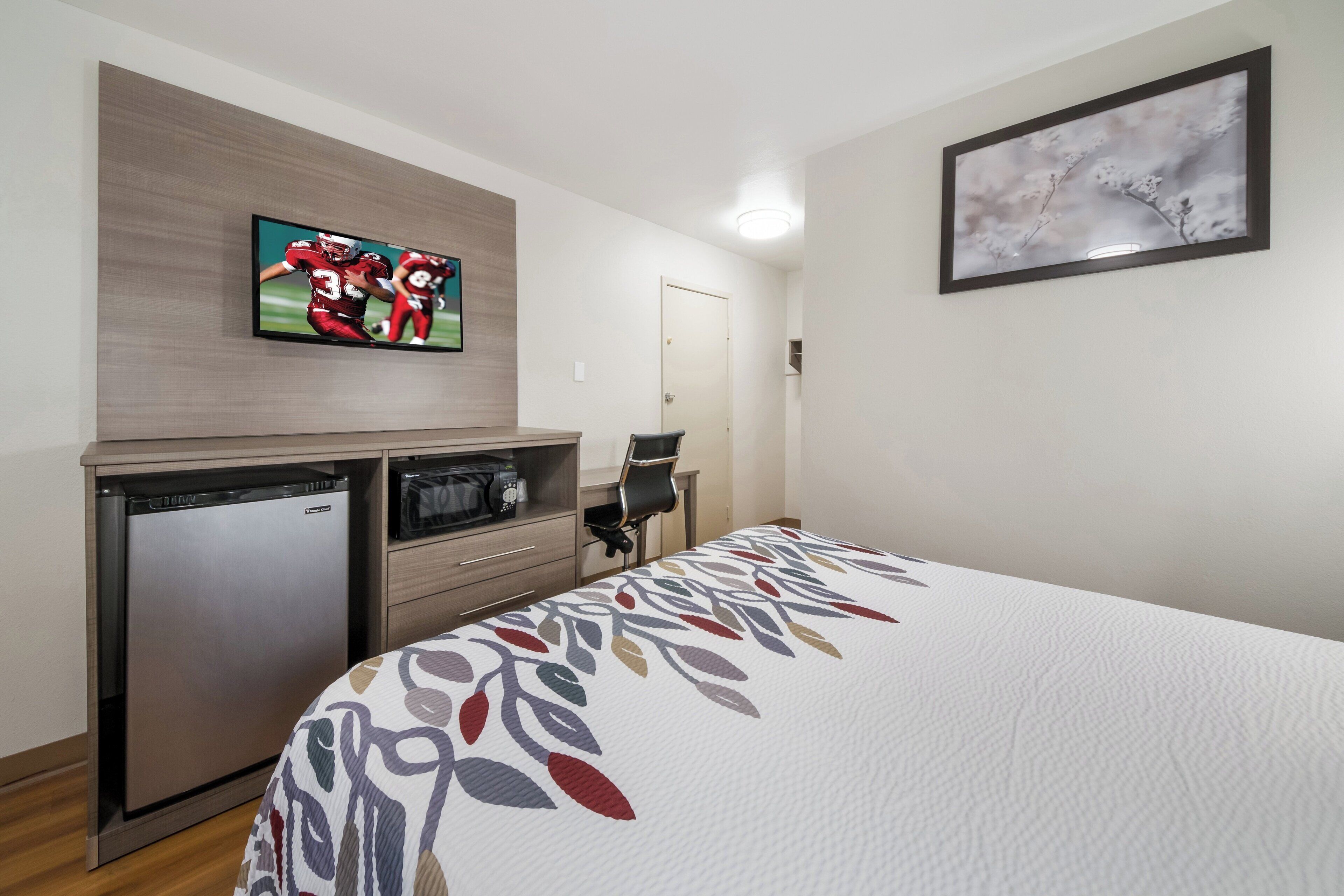 A hotel room with a bed, a television mounted on the wall, a small refrigerator, and a door.
