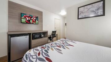 A hotel room with a bed, a television mounted on the wall, a small refrigerator, and a door.