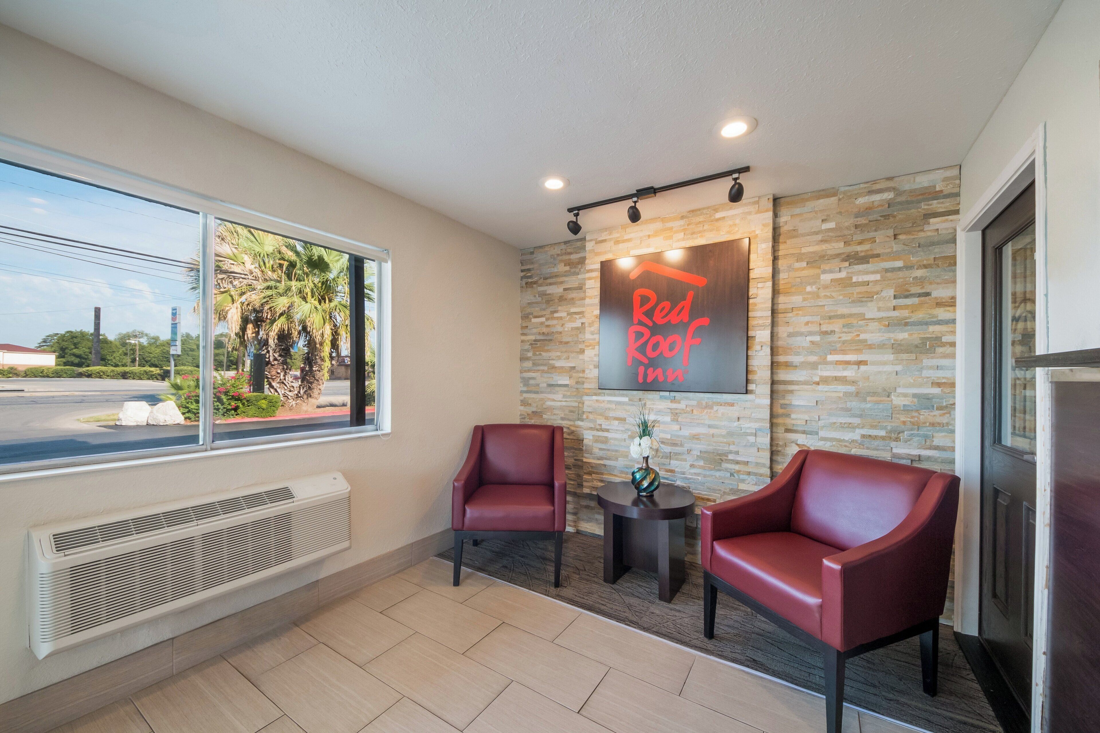 A hotel lobby with two red leather chairs, a stone wall, a Red Roof Inn sign, and a view of palm trees through a large window.