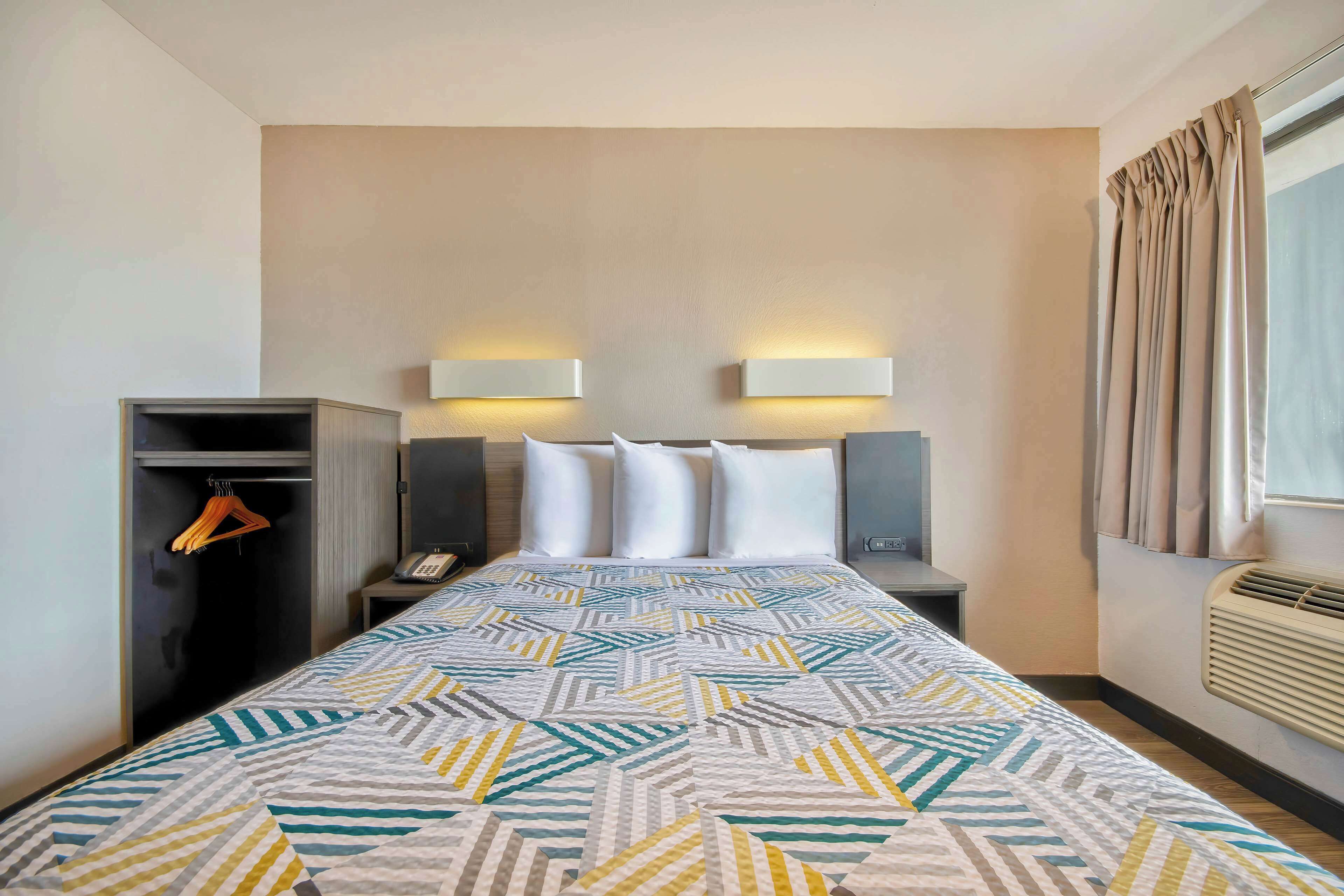Habitación de hotel con cama, armario, mesita de noche, lámpara y ventana con cortinas.