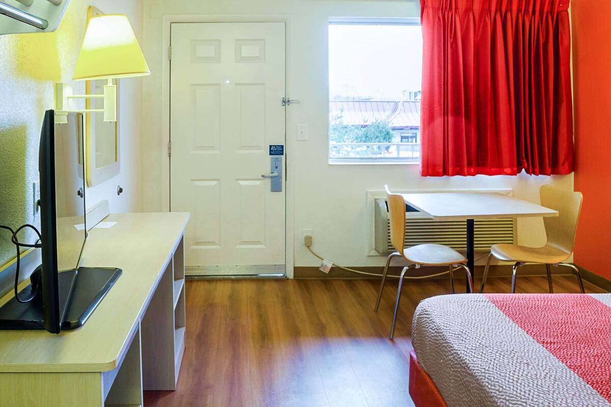 A hotel room with a bed, a desk with a computer, a chair, a window with red curtains, and a door.