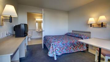 A small, clean room with a bed, a desk, a television, and a bathroom visible through an open door.