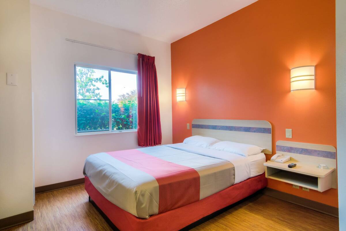 Una habitación de hotel con cama, ventana con vistas a zonas verdes y lámpara fijada a la pared.