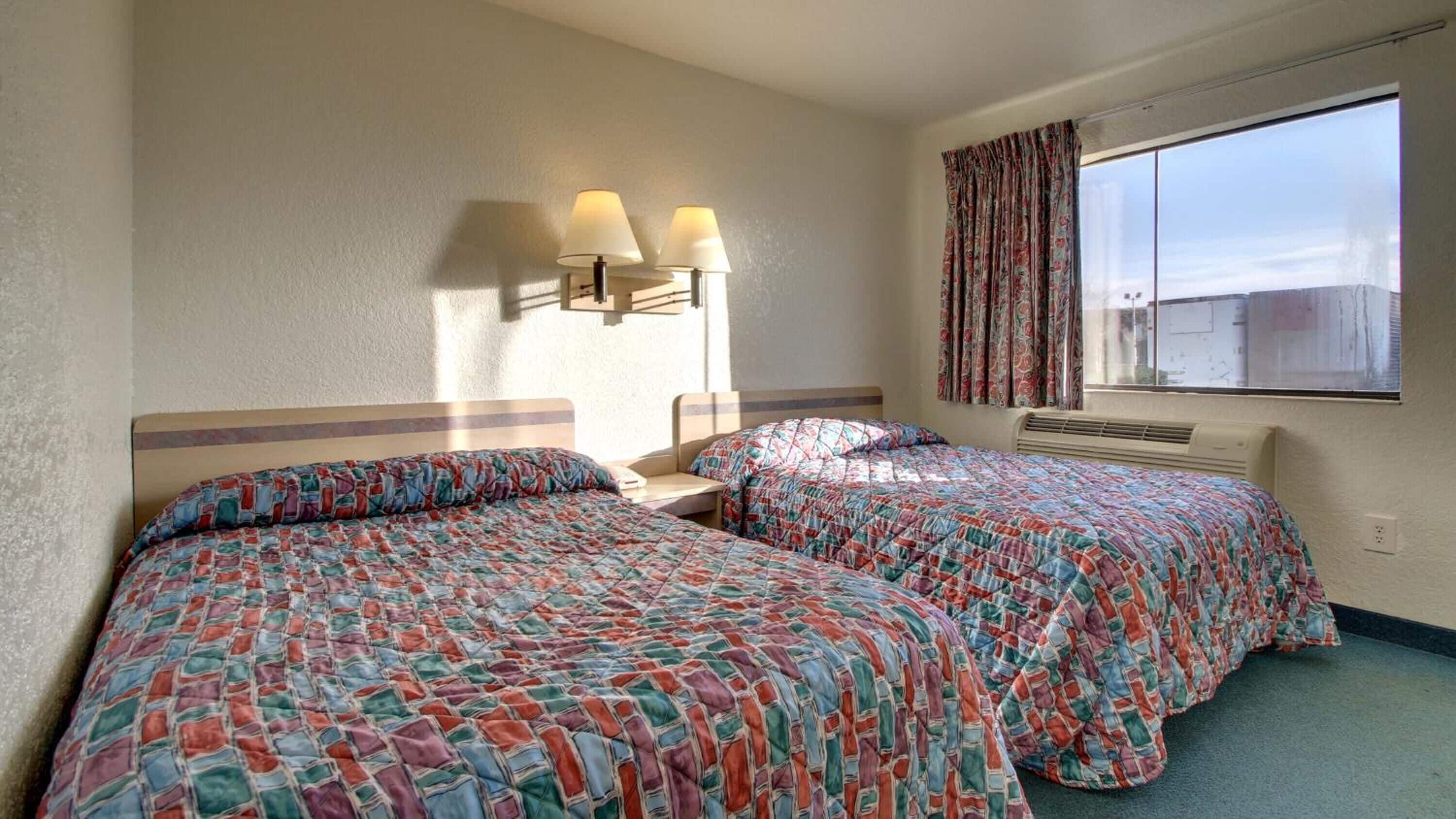 Habitación de hotel con dos camas, una mesita de noche, una lámpara, una ventana con cortinas y un calentador fijado a la pared.