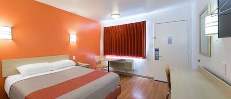 A hotel room with a bed, desk, chair, and an air conditioning unit.
