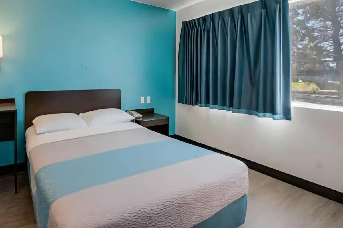 A hotel room with a bed, a chair, a desk, and a window with curtains.