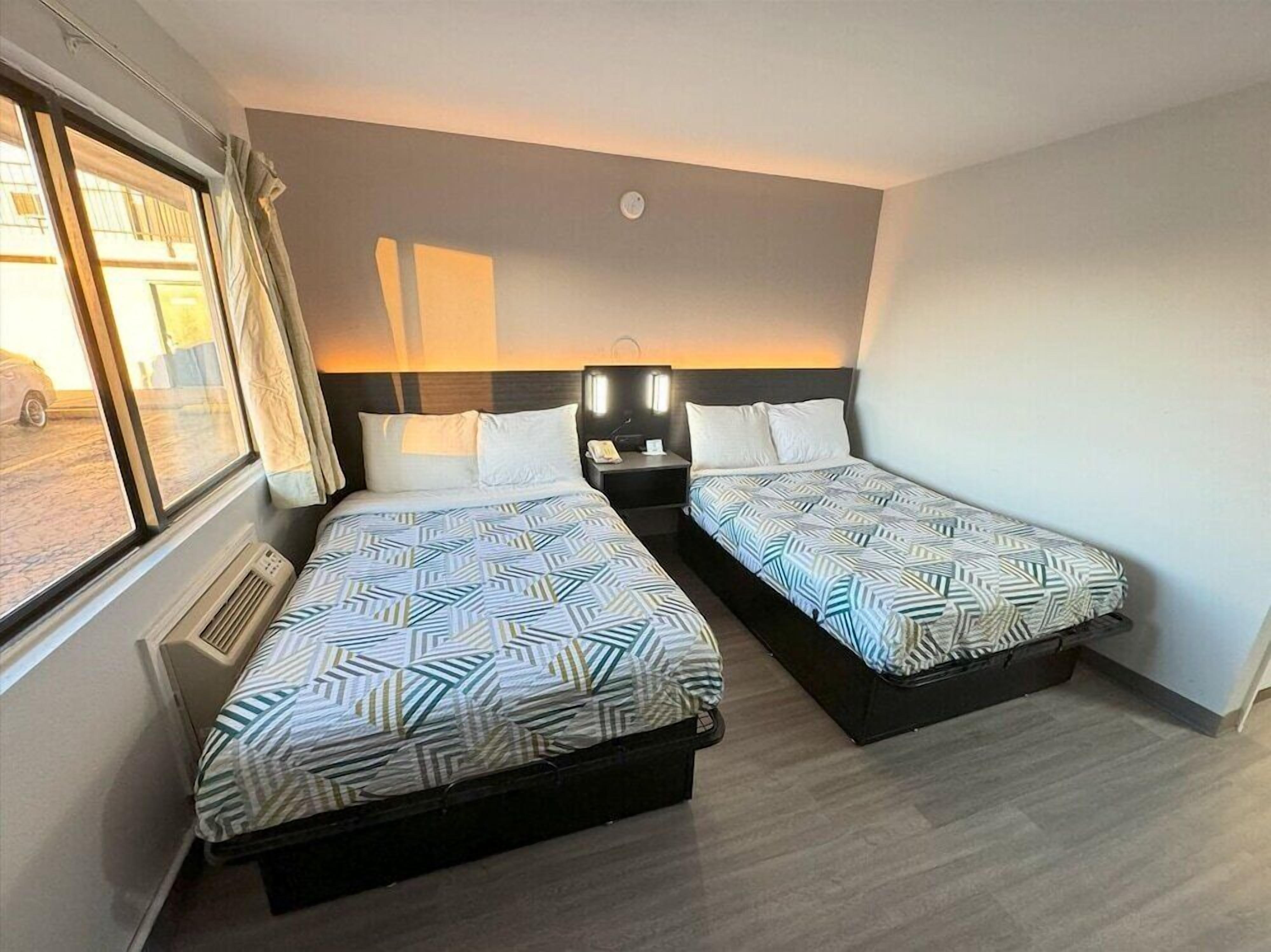 Habitación de hotel con dos camas, una mesita de noche, una lámpara y una ventana con cortinas.