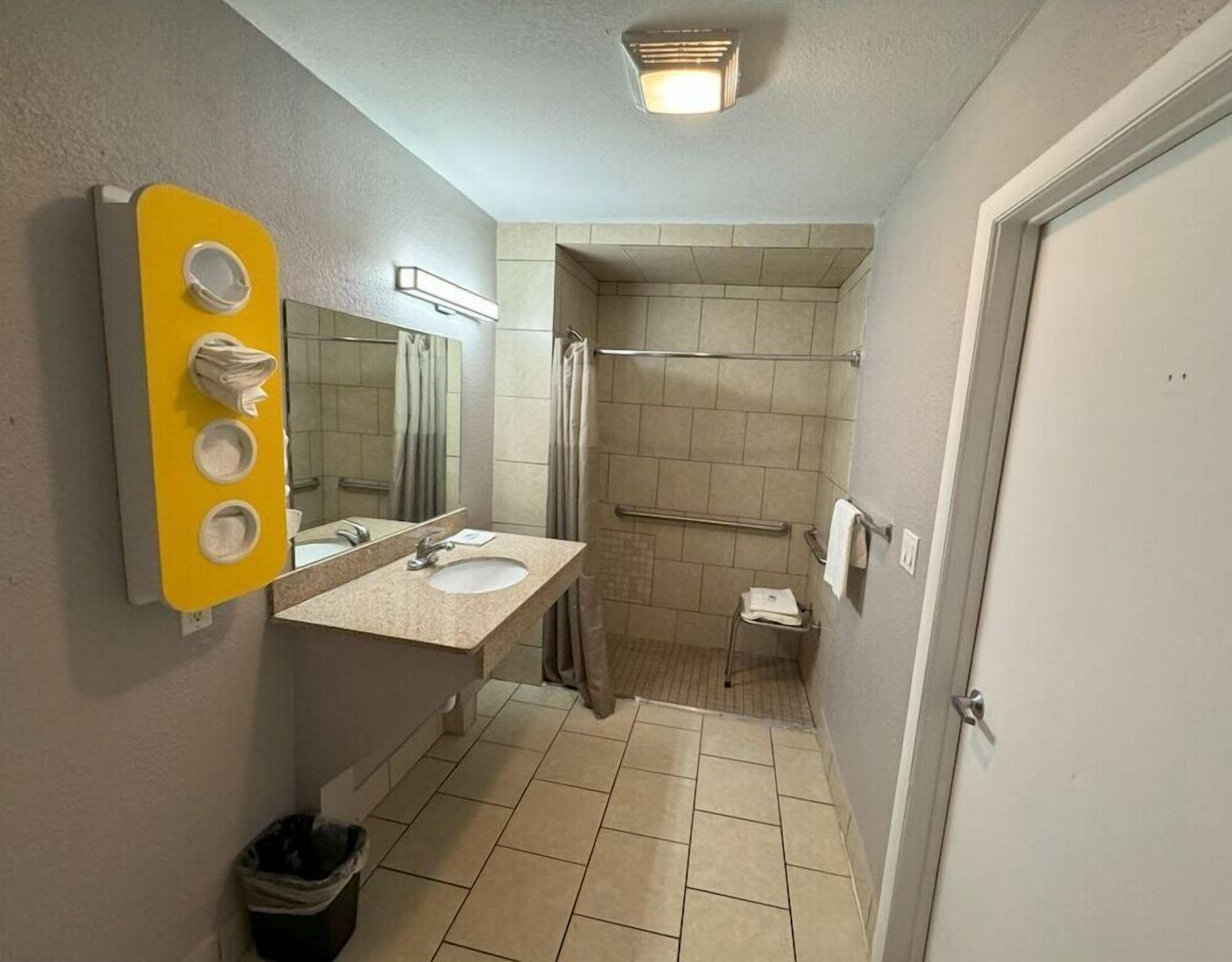A bathroom with a yellow towel dispenser, a sink, a mirror, a shower, and a door.