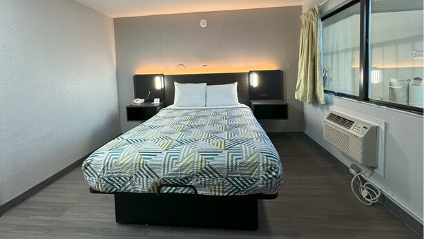 A modern hotel room with a large bed, bedside tables, a window with curtains, and a wall-mounted air conditioner. - Motel 6 Janesville, WI (Janesville)