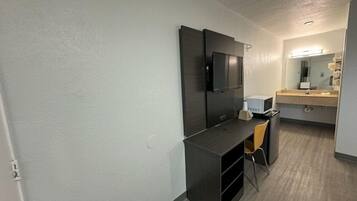 A compact hotel room with a dark wooden desk, a chair, a flat-screen TV mounted on the wall, and a small kitchenette area with a microwave and a sink.