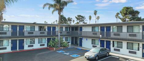 A multi-story motel with blue doors and balconies, a parking lot with marked handicapped spaces, and palm trees in the background.