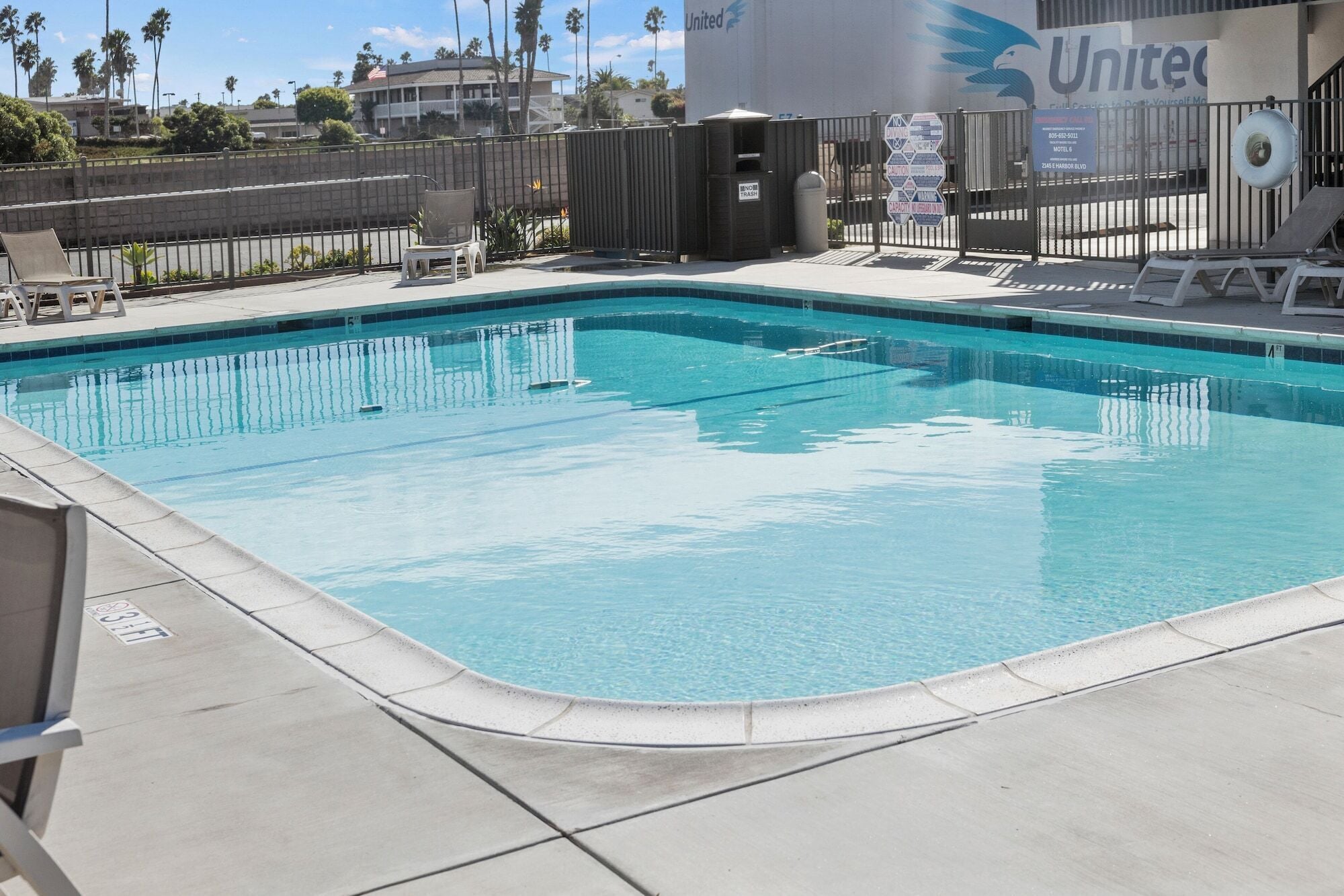 A clear swimming pool with lounge chairs and a trash bin, surrounded by a fence and a building with a 'United' sign.