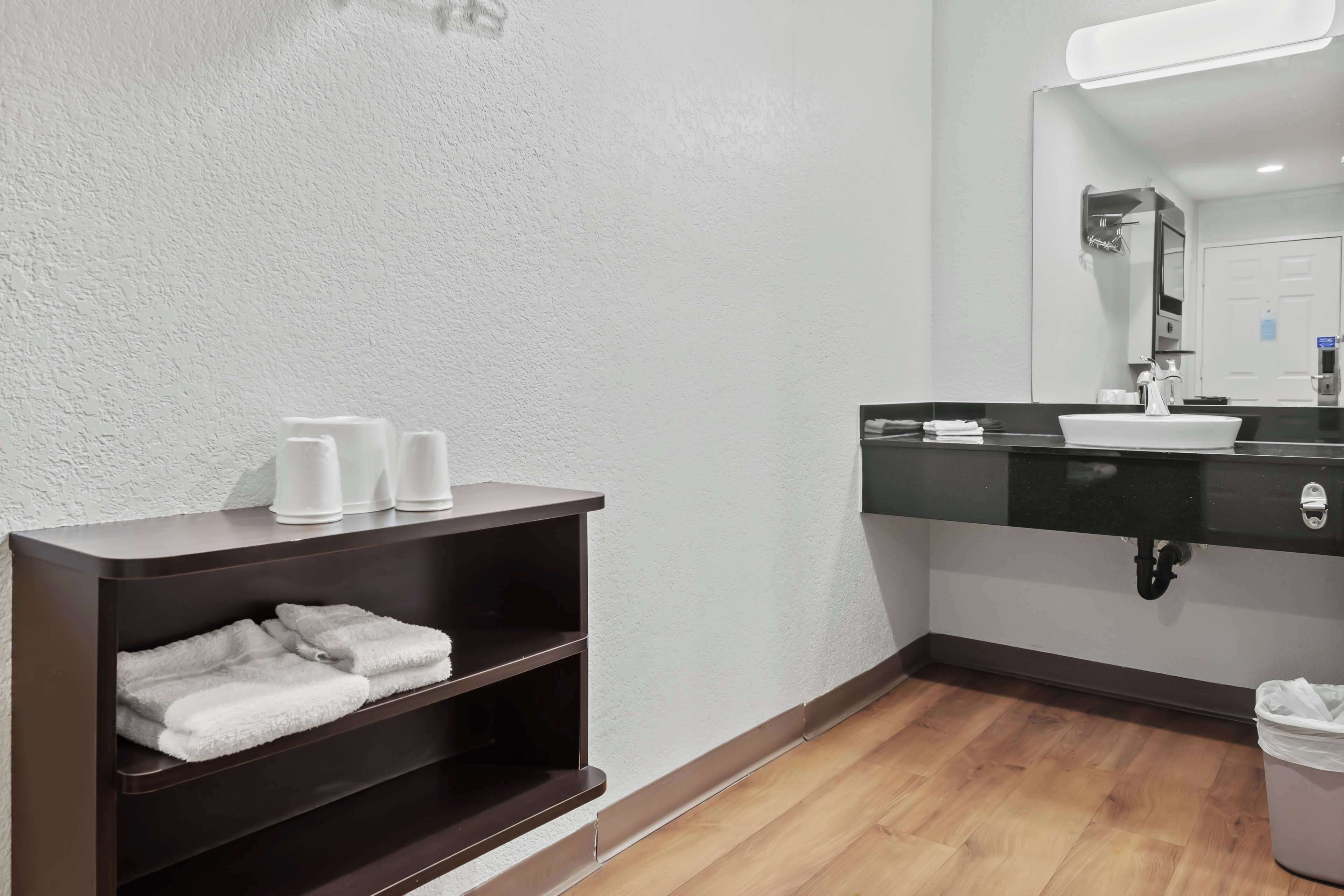 A modern bathroom with a dark wooden vanity, a white sink, and a mirror above it. There are towels on the vanity and a trash can to the right.