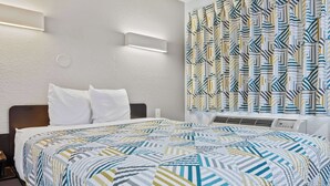 A hotel room with a neatly made bed, a wall-mounted air conditioning unit, and a patterned curtain.