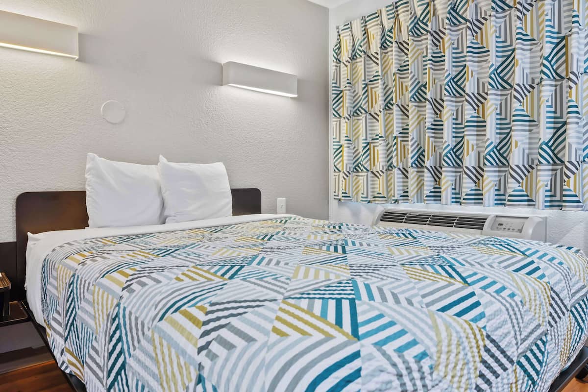 A hotel room with a neatly made bed, a wall-mounted air conditioning unit, and a patterned curtain.