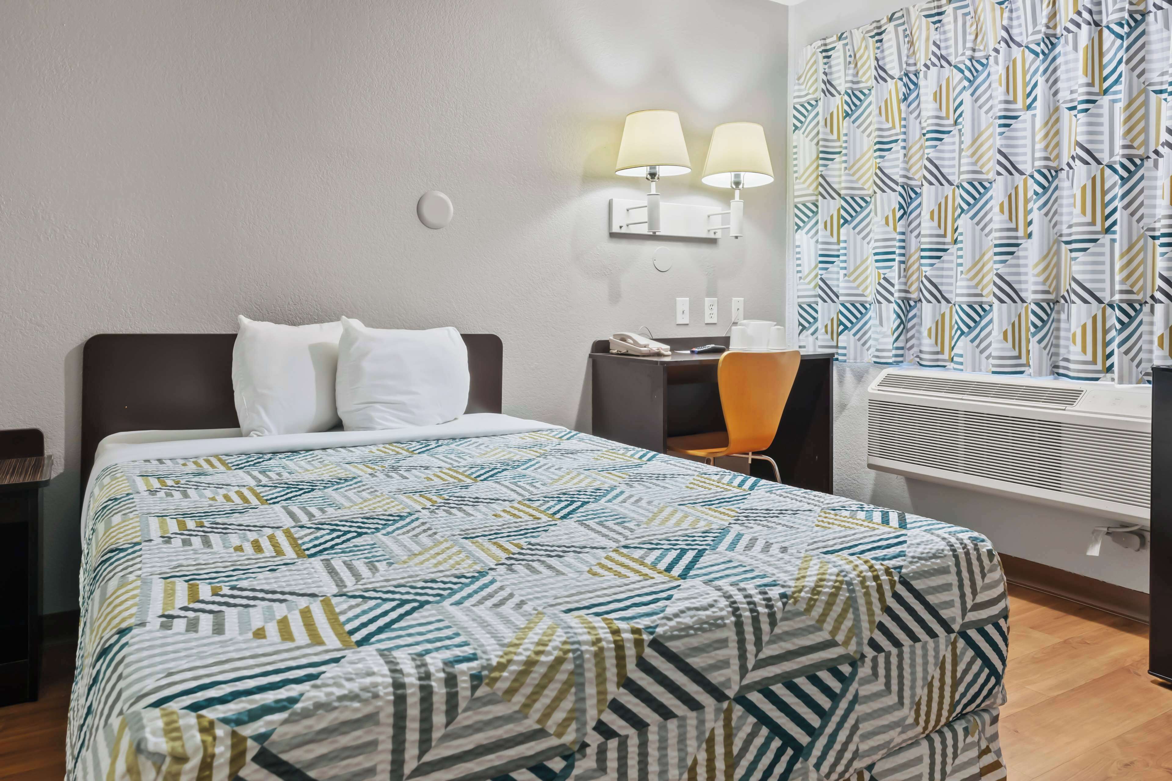 A hotel room with a bed, two bedside lamps, a desk with a phone, and a patterned curtain.