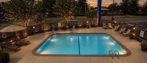 Una piscina rectangular con borde de azulejos, rodeada por un muro de ladrillo bajo. Hay sillas de descanso y una zona de estar cubierta con sombrillas.