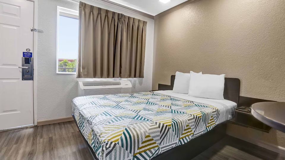 A hotel room with a bed, a window with curtains, an air conditioning unit, a bedside table, and a sink.
