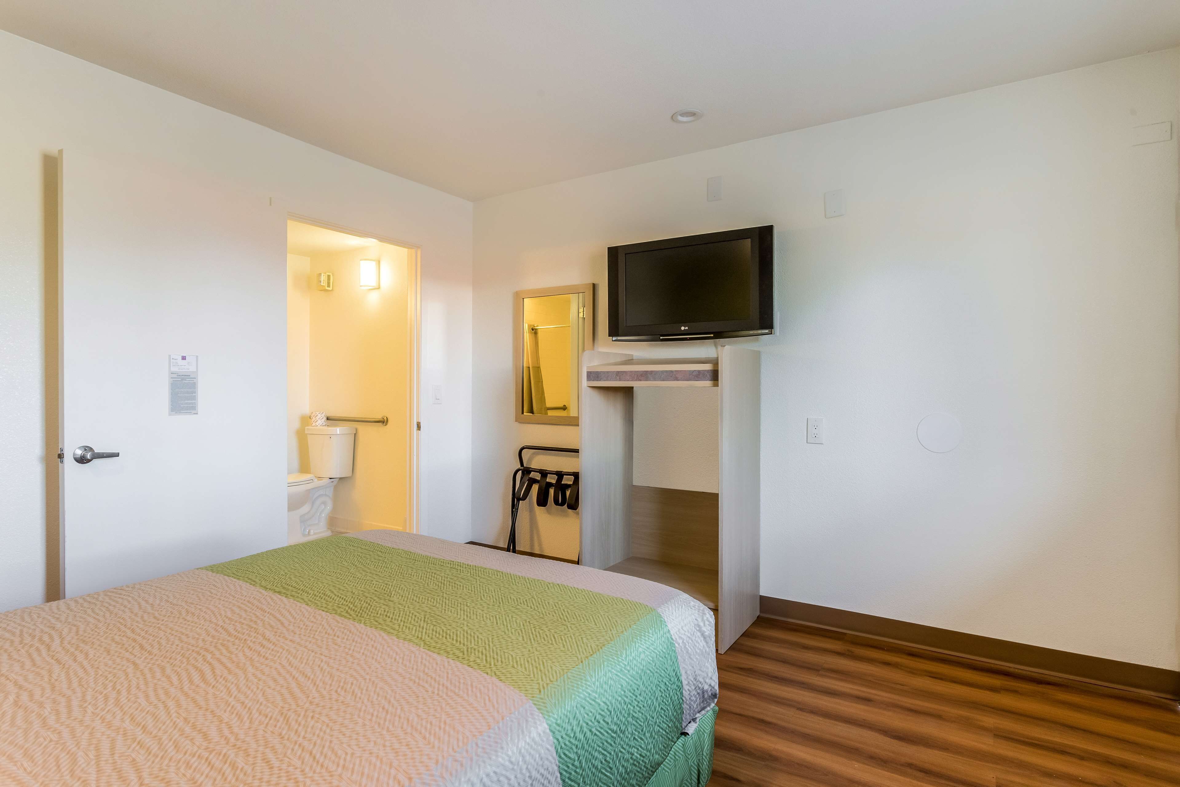 A hotel room with a bed, a television mounted on the wall, a bathroom with a toilet and sink, and a wooden floor.