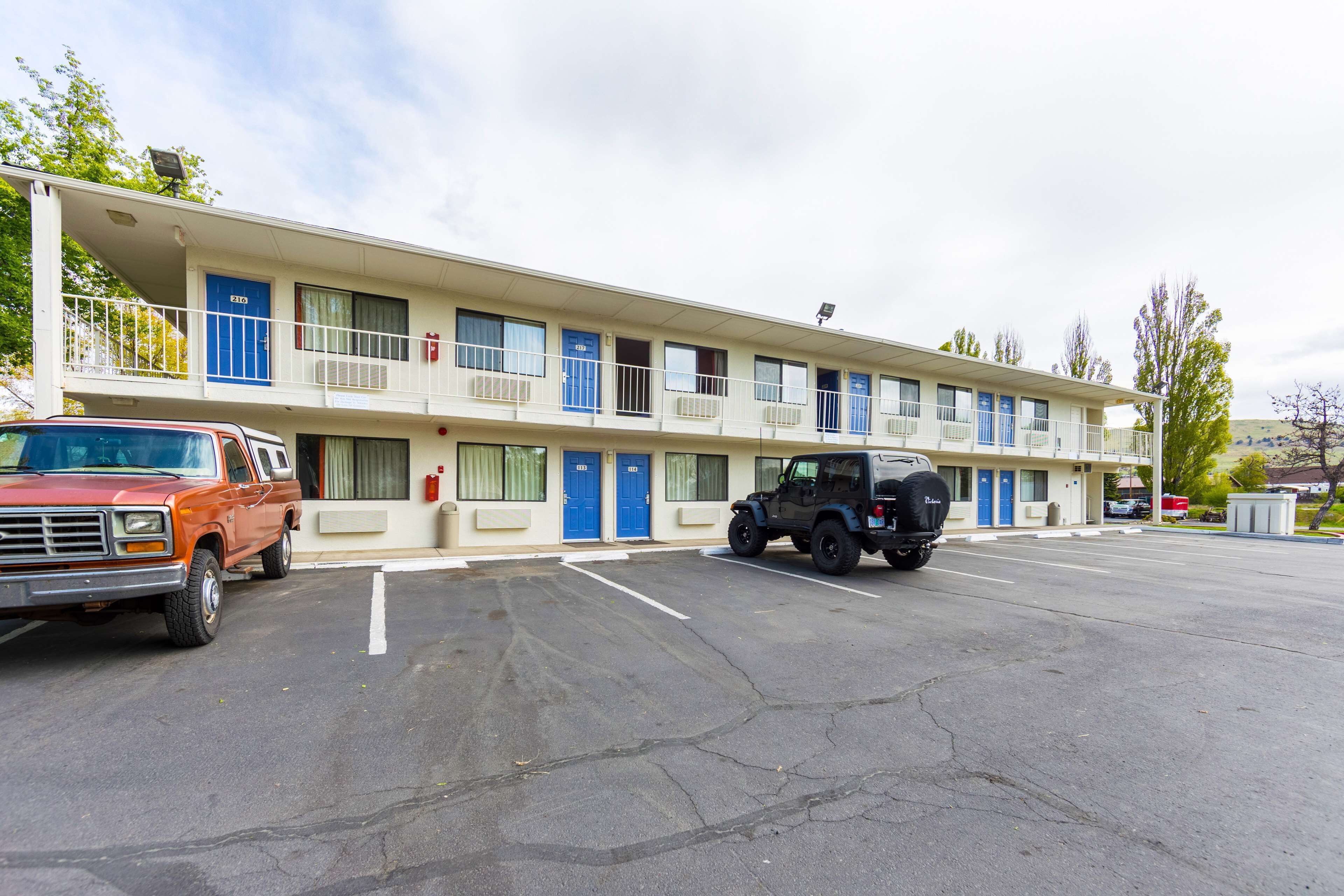 Un motel de dos pisos, con varias habitaciones con puertas azules y un estacionamiento con dos vehículos.