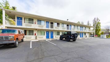 Un motel de dos pisos, con varias habitaciones con puertas azules y un estacionamiento con dos vehículos.