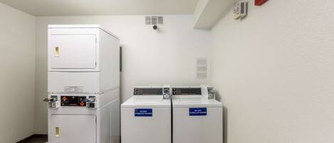 A laundry room with stacked washers and dryers, a fire alarm, and a vent on the ceiling.