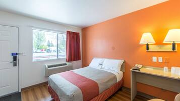 Una habitación de hotel con cama, escritorio, silla, ventana con cortinas y una lámpara de pared.