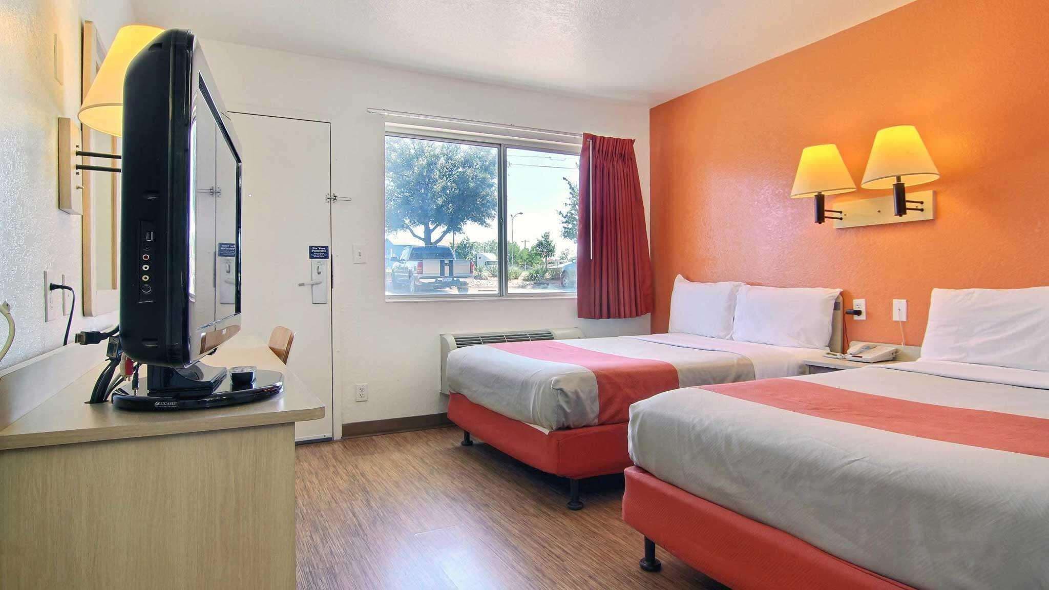 A hotel room with two beds, a desk with a computer, a window with a view of the outside, and a lamp on the wall.