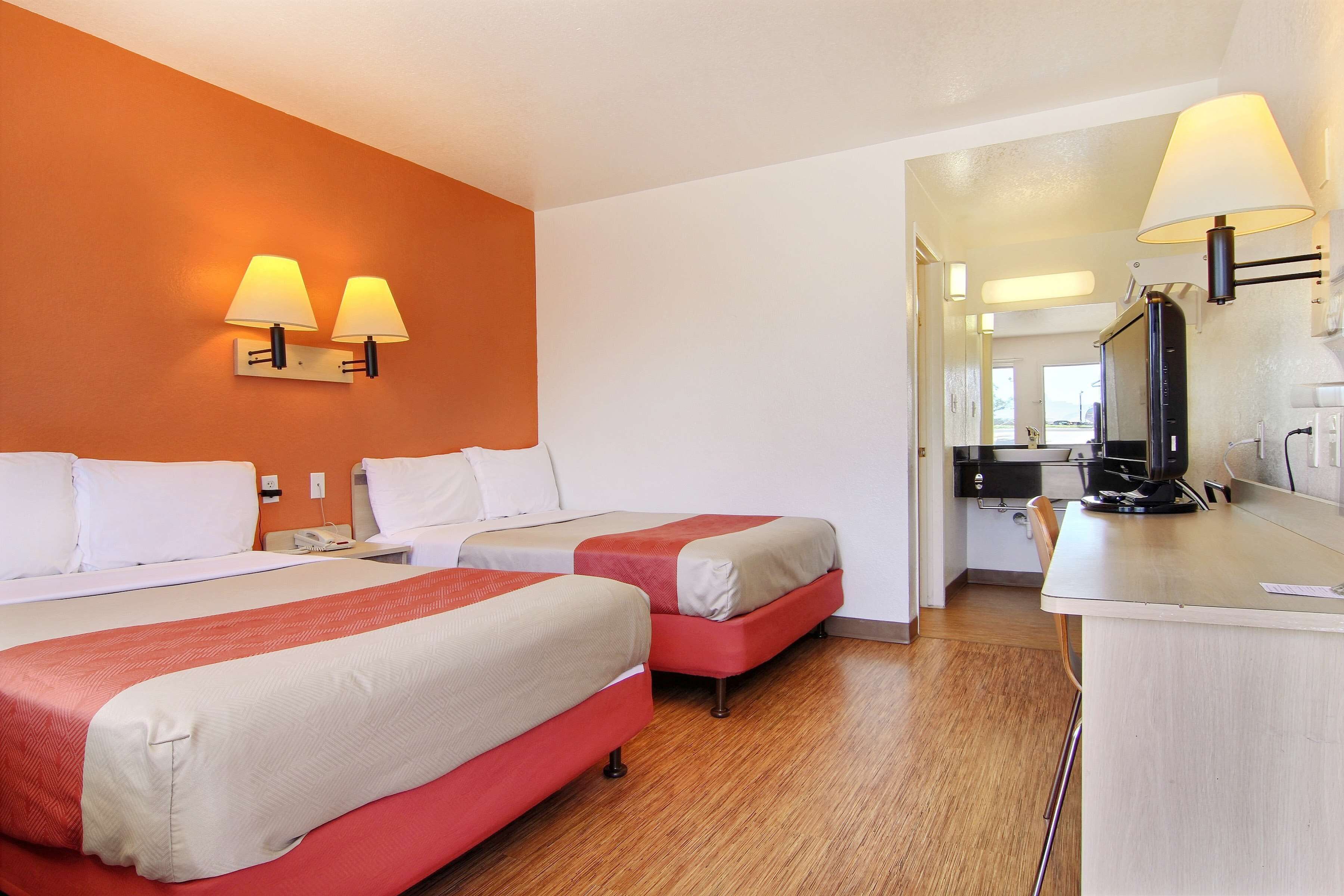 A hotel room with two beds, a desk, a mirror, and a kitchenette.