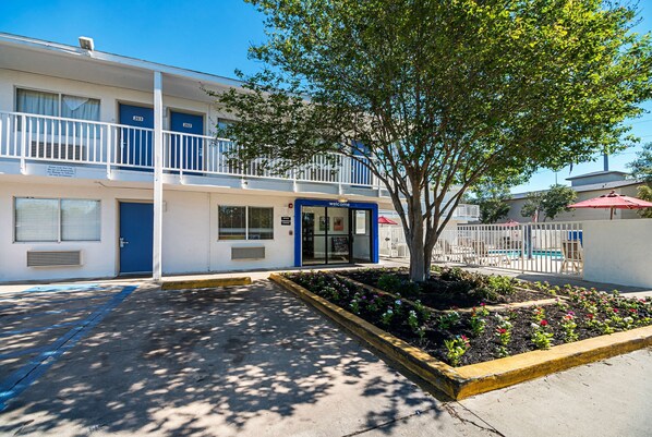 A two-story motel with a blue door marked '203', a small garden with flowers, and a white fence in the background. - Motel 6 Corpus Christi, TX - Northwest (Corpus Christi)