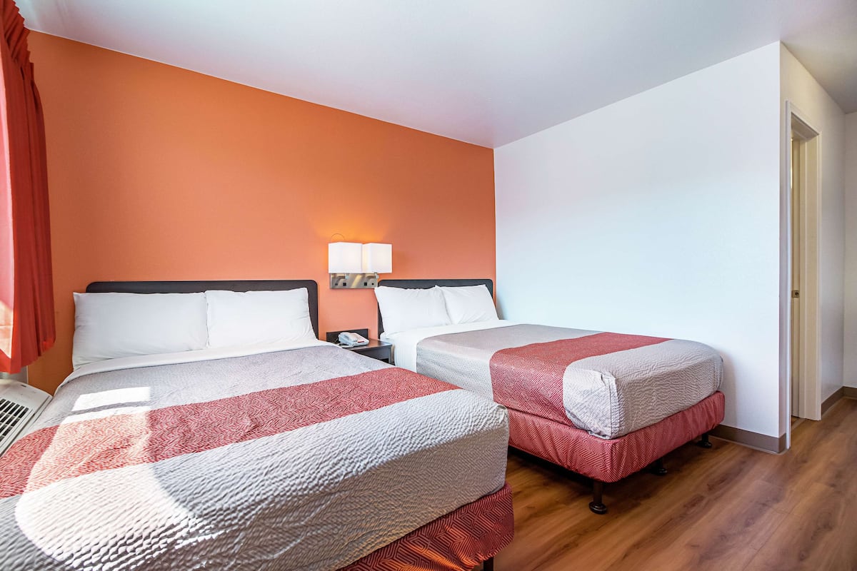 A hotel room with two beds, a nightstand, a lamp, and a window with curtains.