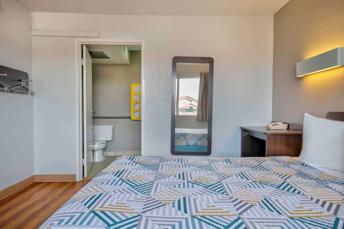 Una habitación de hotel con una cama, una mesita de noche con un teléfono, un espejo y un baño a la vista a través de una puerta abierta.