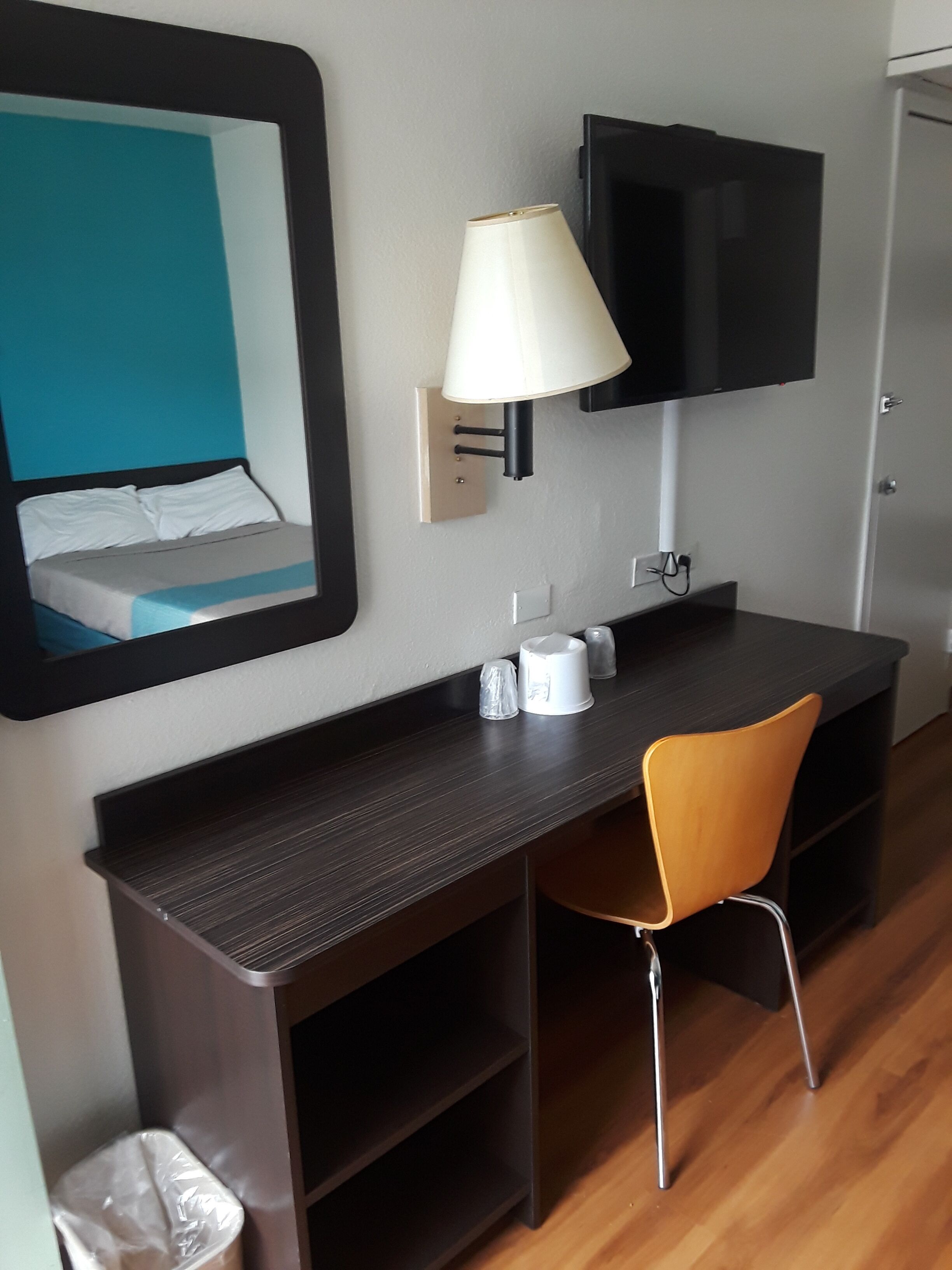 A hotel room with a bed, a desk with a chair, a TV mounted on the wall, and a trash bin.