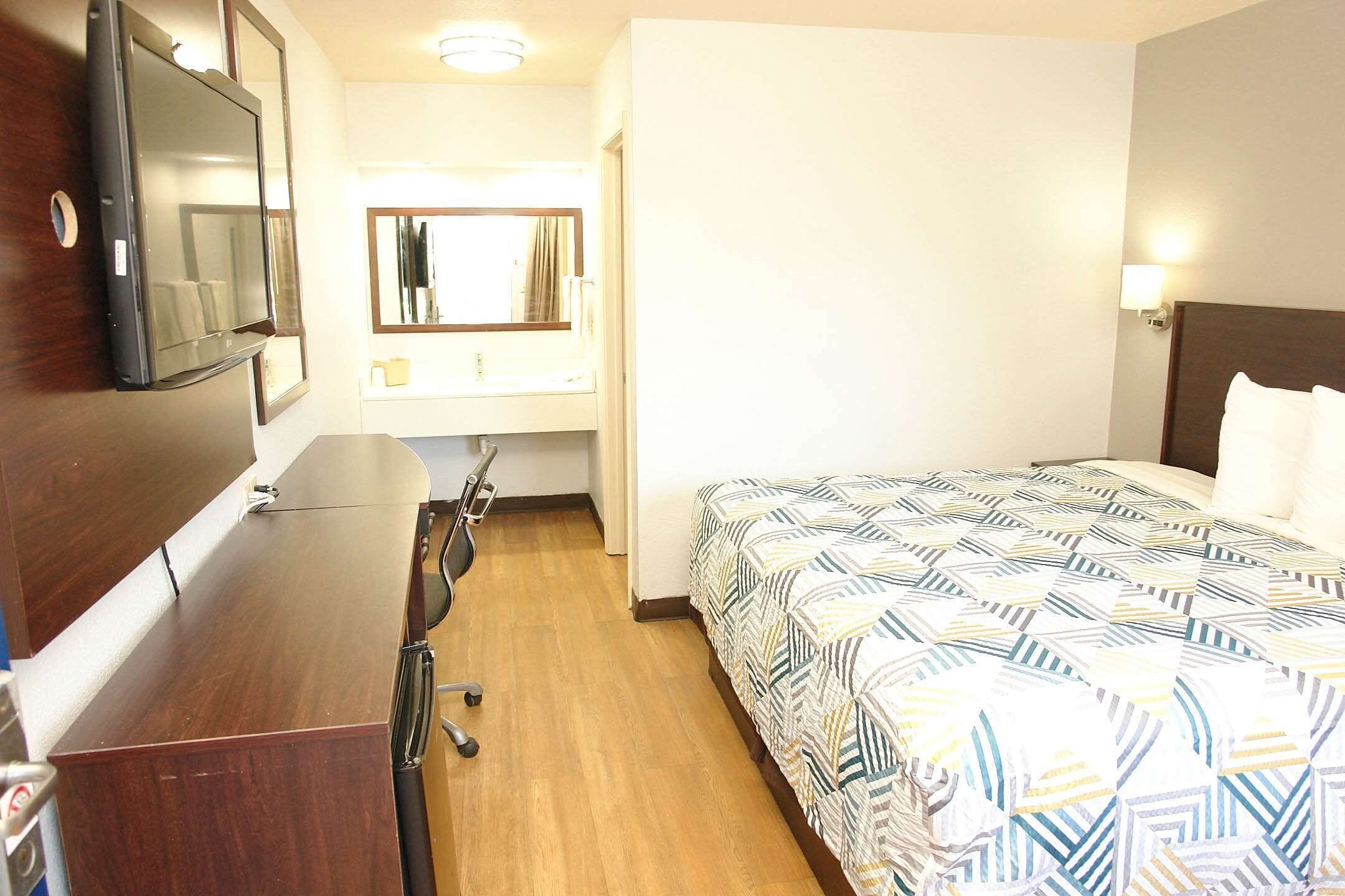 A hotel room with a bed, a desk, a chair, a TV, a mirror, and a nightstand.