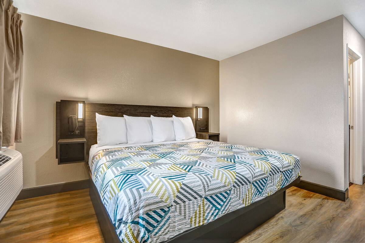 A hotel room with a large bed, a nightstand, a wall-mounted lamp, and a window with curtains.