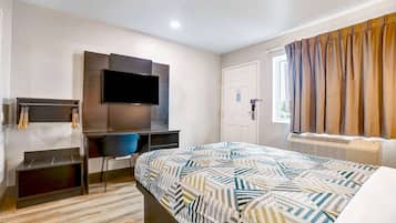 A modern hotel room with a bed, a desk with a flat-screen TV, a chair, and a window with curtains.