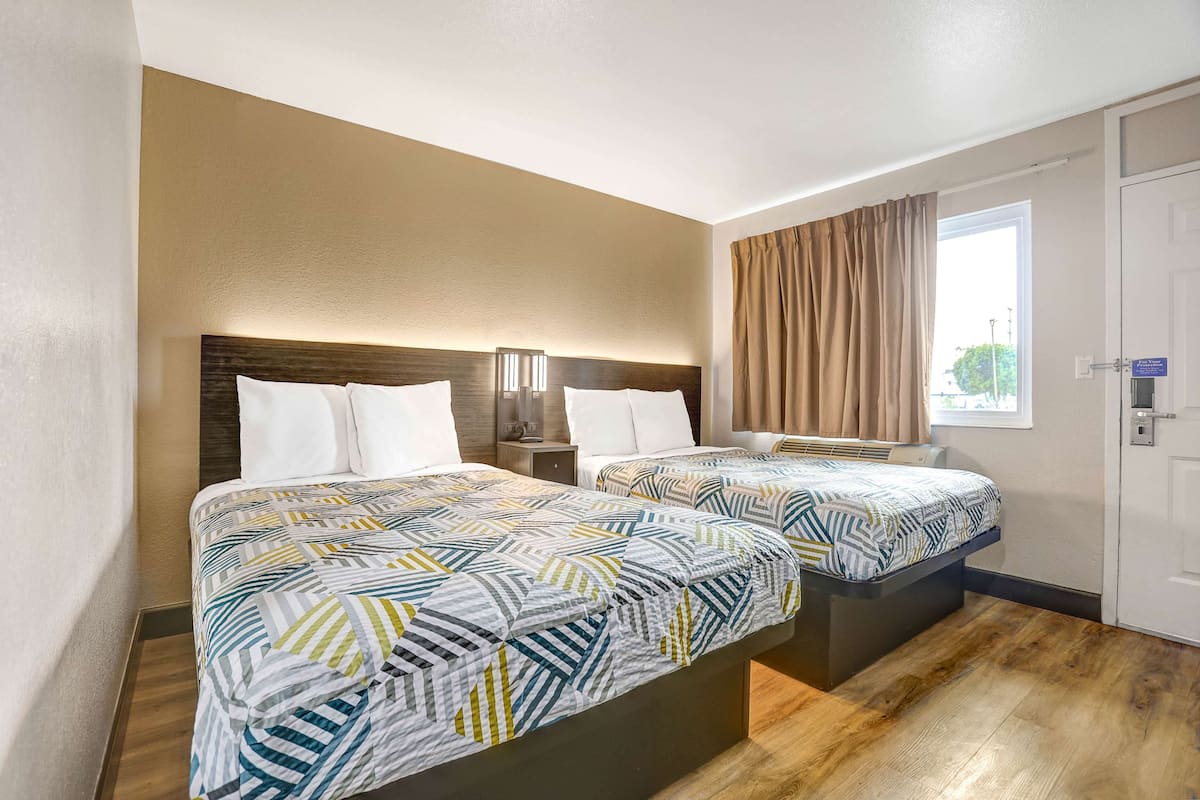 A hotel room with two beds, each with a patterned bedspread, a nightstand with a lamp, and a window with curtains.