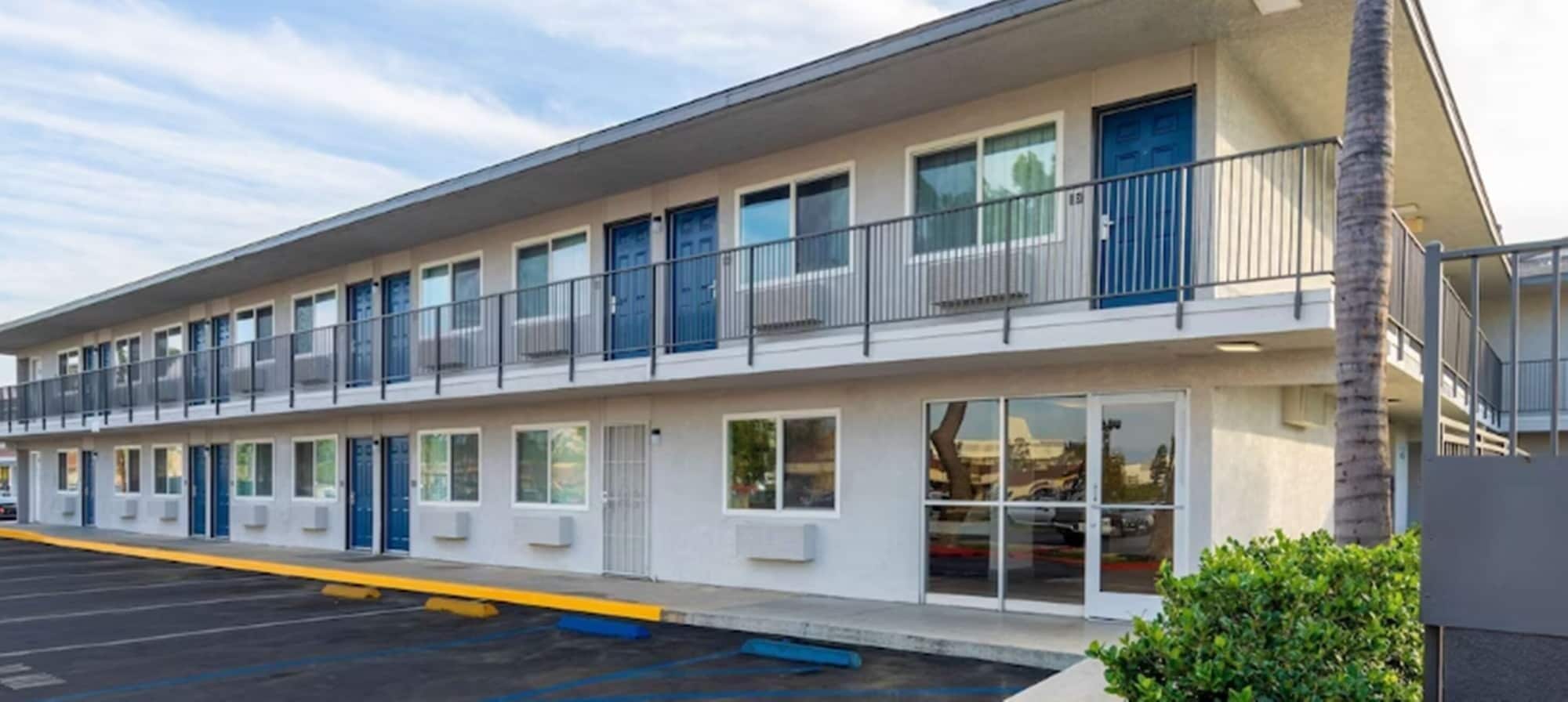 A motel with a row of rooms, a covered parking area, and a palm tree.