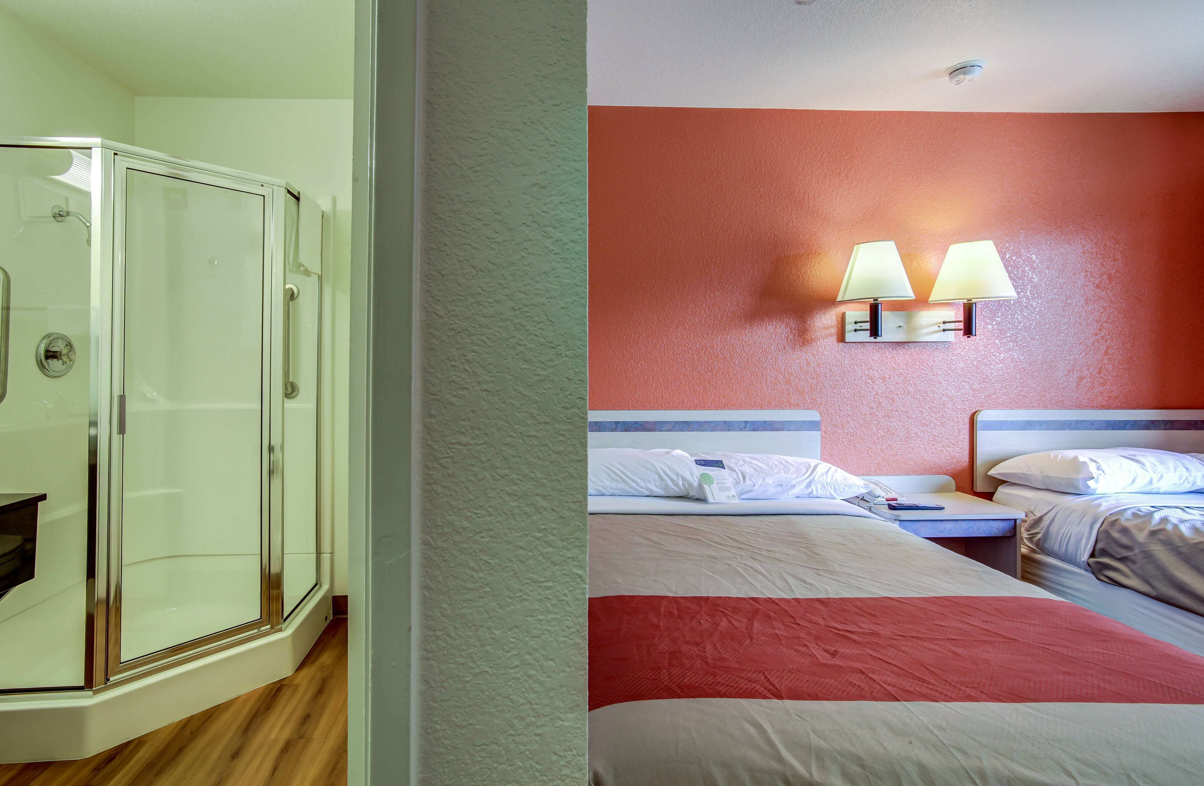 Une chambre d’hôtel dotée d’une douche en verre, de deux lits avec du linge de lit blanc et rouge, et d’une lampe fixée au mur.