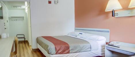 A hotel room with a bed, bedside table, and a desk with a chair. There is a wall-mounted lamp and a small shelf with a telephone.