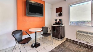 A small room with a white table and two chairs, a television mounted on the wall, a coffee machine, and a window with a view.