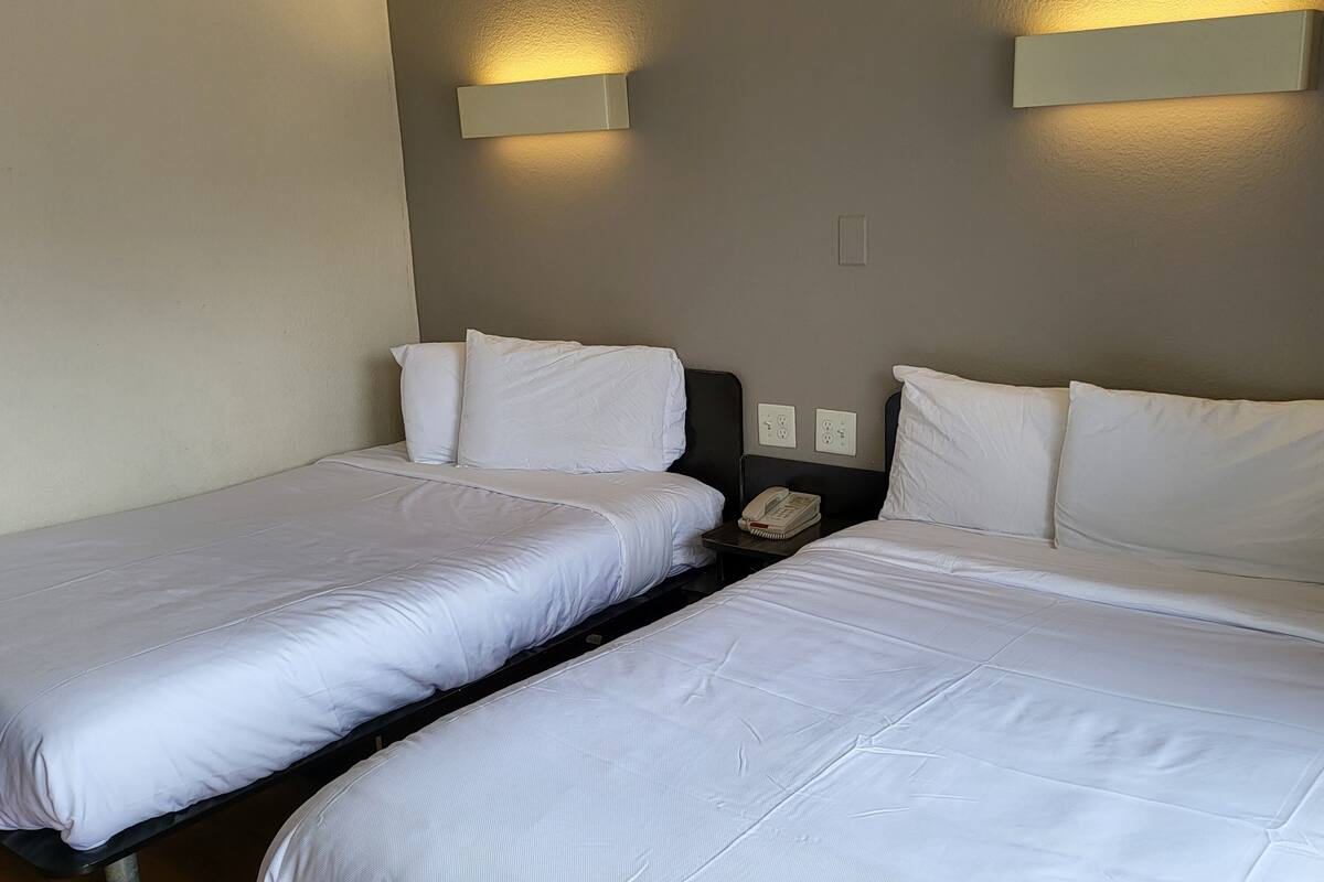 A hotel room with two single beds, each with white bedding and pillows. There are two wall-mounted light fixtures on the wall above the beds.