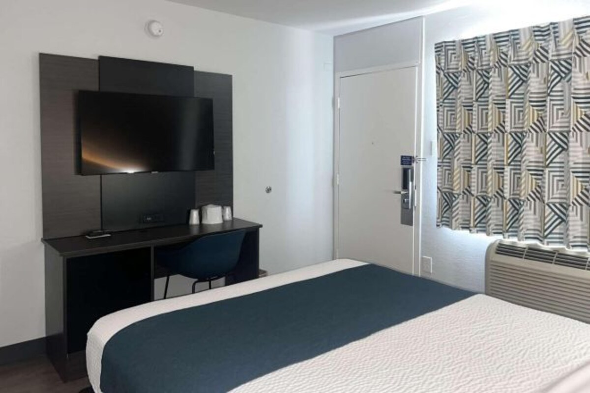 A hotel room with a bed, a desk with a chair, a flat-screen TV mounted on the wall, and a patterned curtain on the window.