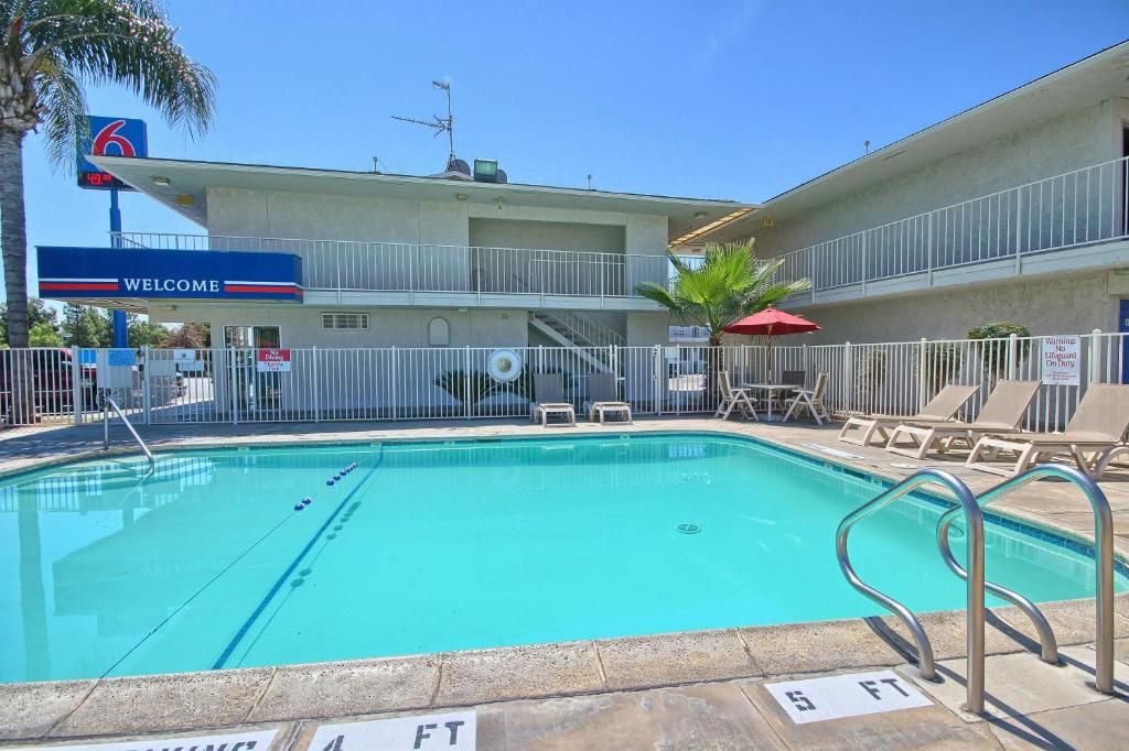 Piscina cercada com área de estar e cadeiras de praia, e um edifício com placa indicando o número 6, característico de um motel.