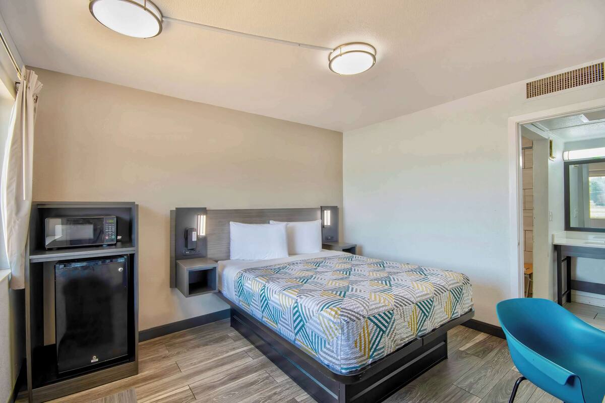 A hotel room with a bed, a microwave, a mini fridge, a nightstand, and a blue chair.