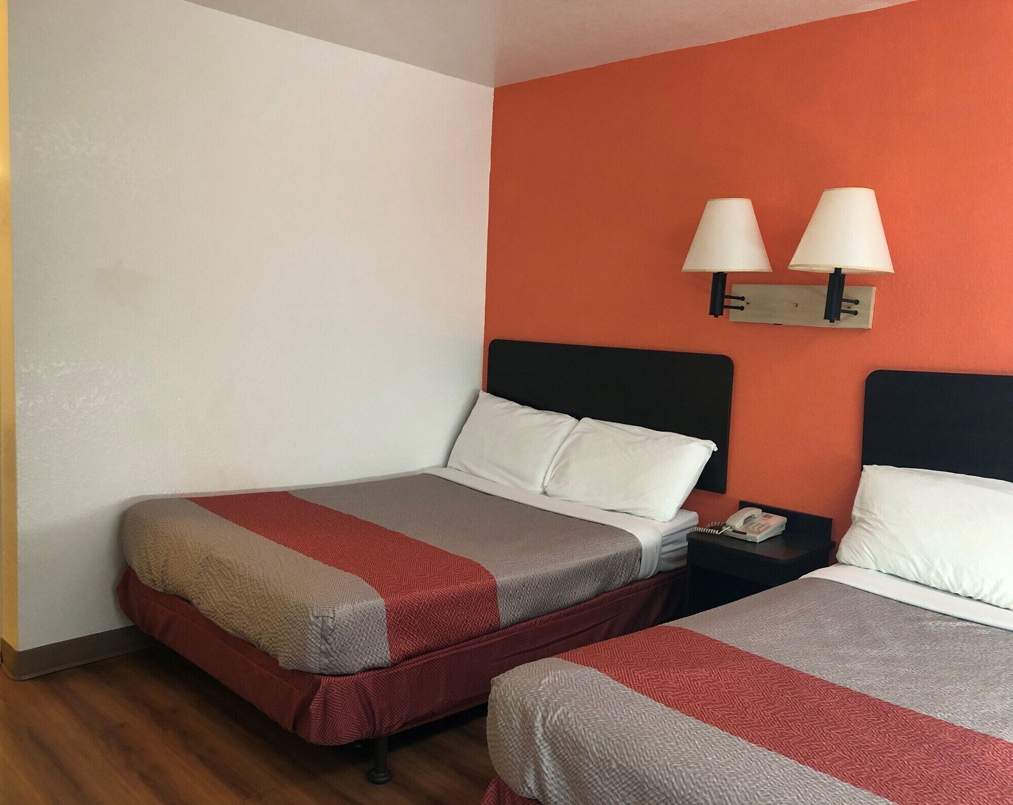 A hotel room with two beds, each with a red and grey bedspread, white pillows, and a black headboard. There are two bedside lamps on the wall, and a telephone on the nightstand.