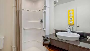Une salle de bain avec des toilettes blanches, un lavabo blanc doté d’un robinet chromé et un distributeur de savon jaune fixé au mur.
