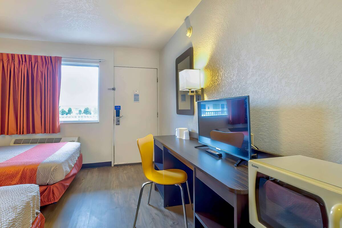 Camera d'albergo con due letti, una scrivania con un computer, una sedia, una finestra con tende e una lampada a parete.